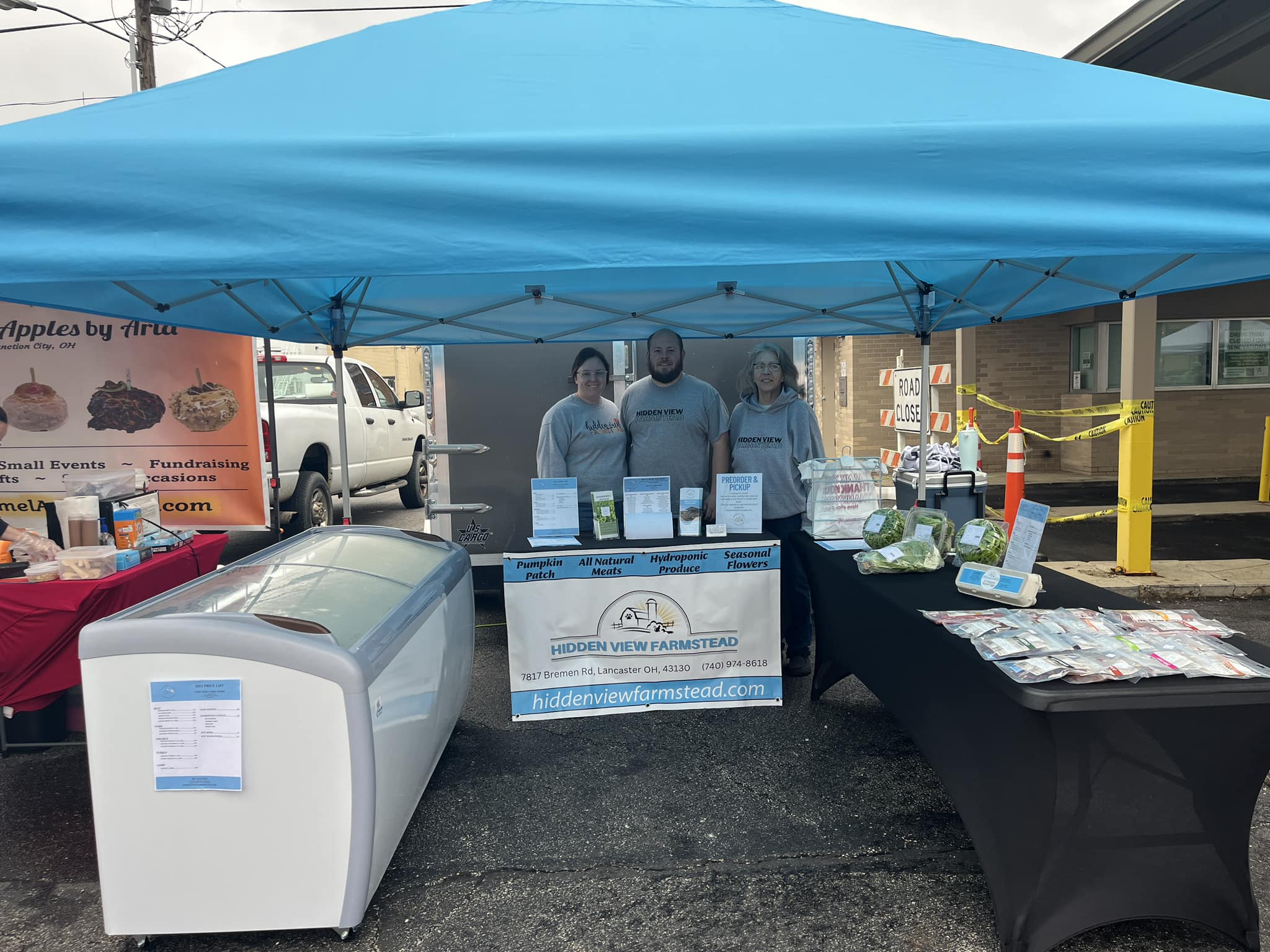 Farmers market booth featuring Hidden View Farmstead, showcasing hydroponic produce, all-natural meats, and seasonal flowers. Three staff members stand behind a table with various products, under a blue canopy. Nearby, a sign indicates the farm's website and contact information.